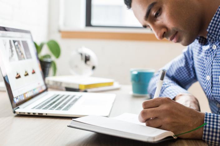 Man writing in notebook with laptop in background